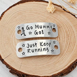 May include: Two rectangular silver-toned metal shoe tags with rounded corners and holes on each side. The top tag features a runner icon and the text "Go Mummy Go!" with stars. The bottom tag features the text "Just Keep Running" with stars.