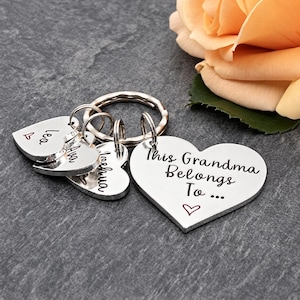 Pode incluir: Um porta-chaves de metal prateado com um pendente grande em forma de cora&ccedil;&atilde;o gravado com o texto This Grandma Belongs To... e tr&ecirc;s pendentes mais pequenos em forma de cora&ccedil;&atilde;o com os nomes Lea, Ava e Joshua, cada um com um pequeno cora&ccedil;&atilde;o.