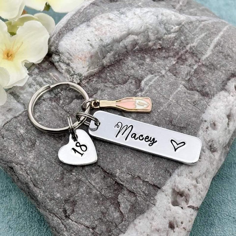 May include: Silver keyring with a heart charm engraved with the number 18, a small champagne bottle charm, and a rectangular tag engraved with the name "Macey" and a heart.