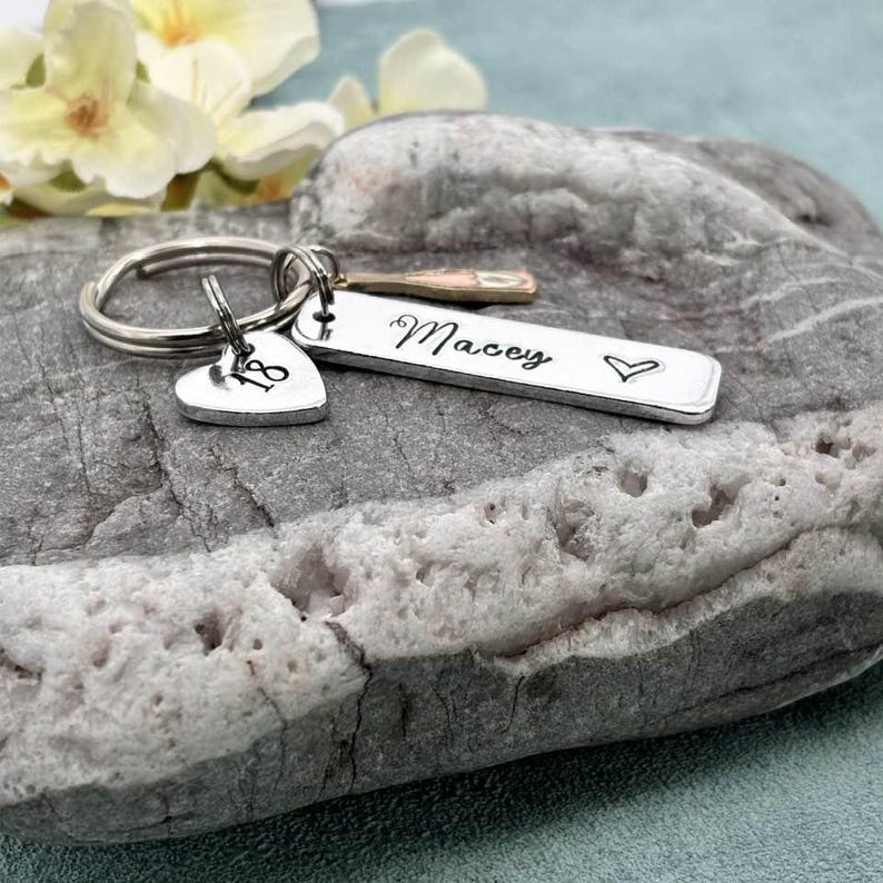 May include: Silver keyring with a heart charm engraved with the number 18 and a bar charm engraved with the name "Macey" and a heart.