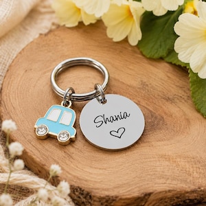 May include: A silver-toned metal keyring featuring a circular disc engraved with the name Shania and a small heart symbol, accompanied by a light blue car-shaped charm with two clear crystal accents set into the wheels of the vehicle.