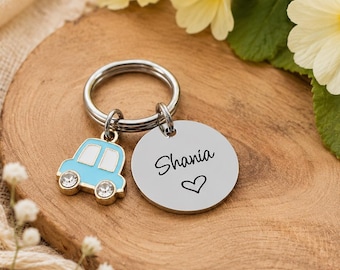 May include: A silver-toned metal keyring featuring a circular disc engraved with the name Shania and a small heart symbol, accompanied by a light blue car-shaped charm with two clear crystal accents set into the wheels of the vehicle.