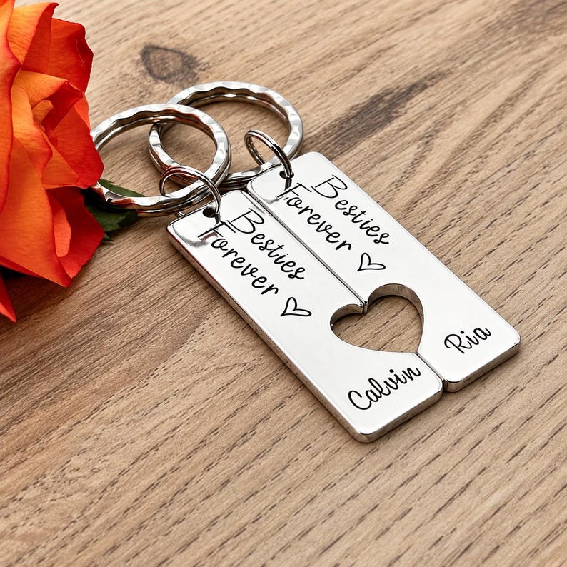 May include: Two silver-toned rectangular metal keychains with split rings. Each piece features the engraved text Besties Forever, a heart outline, and the names Calvin and Ria. When placed together, the cutouts form a single heart shape.