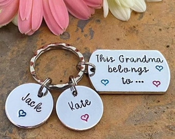 May include: A silver-toned metal keychain featuring a rectangular tag engraved with the text "This Grandma belongs to..." and two circular charms engraved with the names "Jack" and "Kate," each accented with small blue and pink heart symbols.