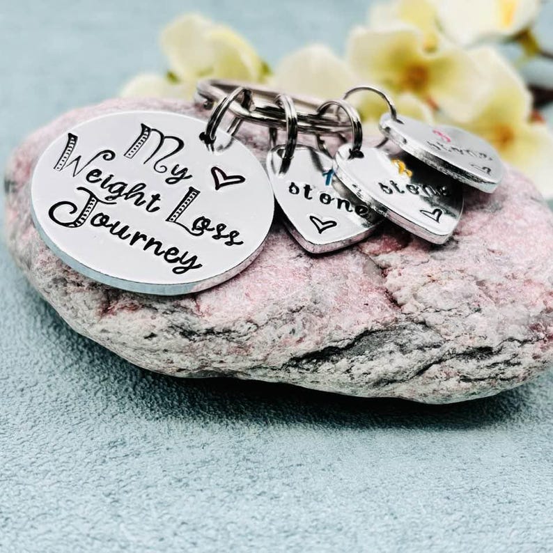 May include: A silver-toned metal keychain featuring a large circular disc engraved with the text My Weight Loss Journey and a small heart symbol. Attached are three smaller heart-shaped charms, each engraved with the word stone and a heart.