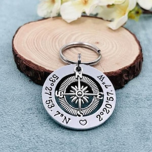 May include: A circular silver-toned metal keychain featuring a central compass rose design. The outer edge is engraved with the coordinates 53° 27' 53.7" N and 2° 20' 57.7" W, accompanied by a small heart symbol at the bottom of the circular charm.