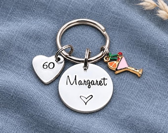 May include: A silver-toned metal keychain featuring a circular ring, a heart-shaped charm engraved with the number 60, a round disc charm engraved with the name Margaret and a heart symbol, and a small colorful cocktail glass charm.