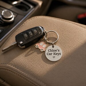 Puede incluir: Un llavero de coche negro unido a una anilla de metal plateado con dos dijes: un coche peque&ntilde;o de color rosa con ventanas blancas y un disco redondo de plata grabado con el texto Chloe's Car Keys y un peque&ntilde;o s&iacute;mbolo de coraz&oacute;n debajo.