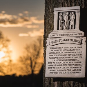 May include: A white stone plaque attached to a tree trunk. The plaque has the words "Tomb of the Unknown Soldier" and "Never Forget Garden" on it. The plaque features a relief sculpture of three figures in togas. The text on the plaque reads: "This garden is a living tribute to all of America's veterans and their families. In silence and respect, this is a place to remember why millions of Americans have fought and died for our liberty and our freedom. Here we renew our promise to fulfill America's sacred duty to never forget. Here we renew our mutual pledge to support them with: "Our lives, our fortunes, and our sacred honor."