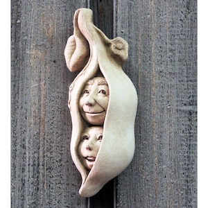 May include: A white ceramic sculpture of two smiling faces peeking out of a pea pod.