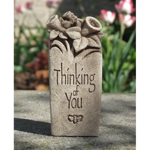 May include: A small, rectangular, light brown stone garden marker with the words "Thinking of You" carved into it. The marker is decorated with a carved floral design and a small bee.