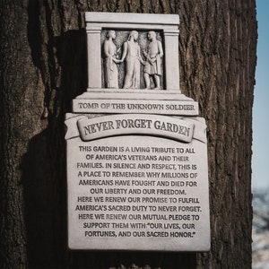 May include: A white stone monument inscribed with the words "Tomb of the Unknown Soldier" and "Never Forget Garden". The monument features a carved image of three figures and a plaque with a message about remembering veterans and their families.