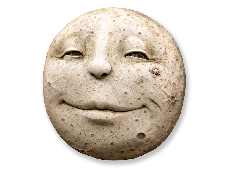 Op de afbeelding: Een ronde, gebroken witte sculptuur van een lachend maangezicht. De maan heeft een textuur met kraters en gegraveerde lijnen. Een kleine, bloemachtige versiering bevindt zich op de rechterwang. De ogen van de maan zijn lichtjes gesloten.