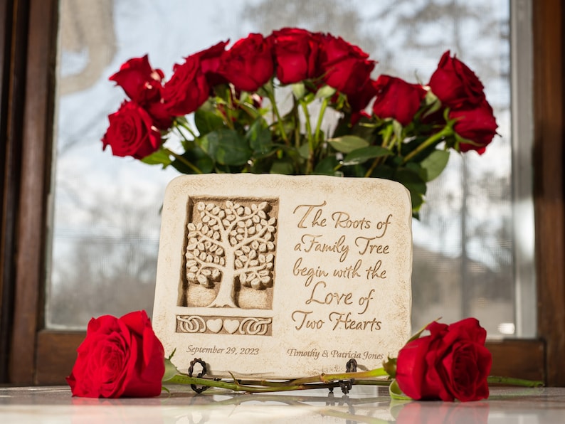 May include: A white stone plaque with a carved tree design and the inscription "The Roots of a Family Tree begin with the Love of Two Hearts". The plaque is decorated with a heart and a Celtic knot. The date "September 29, 2023" and the names "Timothy & Patricia Jones" are also inscribed on the plaque. The plaque is surrounded by red roses.