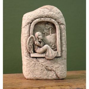 May include: A stone sculpture of an angel with wings sitting in a window looking out at a landscape with a tree. The sculpture is painted in shades of gray and white.