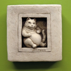 May include: A white ceramic wall plaque with a raised image of a cat sitting in a window. The cat is white and has a round belly. The window frame is also white and has the word "relax" written below the cat.