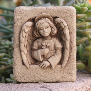 May include: A stone sculpture of an angel with wings holding a bouquet of flowers. The sculpture is framed in a square stone border.