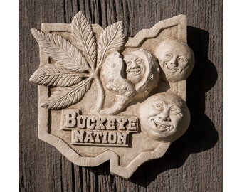 May include: A white ceramic wall plaque shaped like the state of Ohio with a buckeye nut and leaves, and three smiling faces. The plaque says "Buckeye Nation".