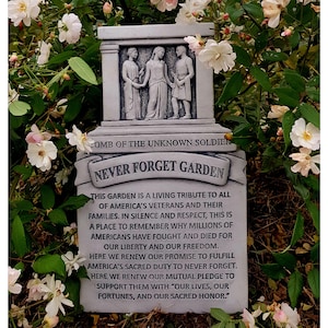 May include: A grey stone memorial with the words "Never Forget Garden" and a quote about honouring veterans and their families. The memorial features a sculpted image of three figures, likely representing soldiers.