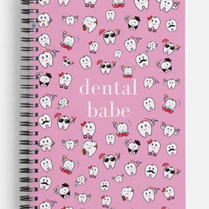K&ouml;nnte beinhalten: Ein rosa Notizbuch mit Spiralbindung, wei&szlig;em Hintergrund und einem Muster aus Cartoon-Z&auml;hnen mit verschiedenen Gesichtsausdr&uuml;cken. Der Text "dental babe" ist in wei&szlig; in der Mitte des Notizbuchs gedruckt.