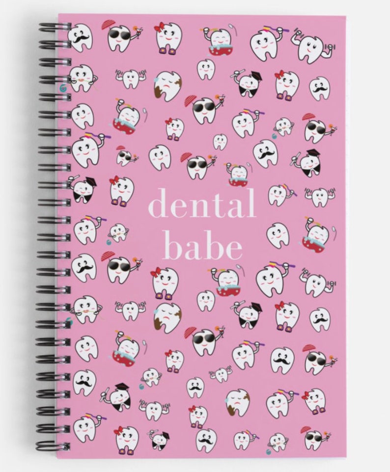 K&ouml;nnte beinhalten: Ein rosa Notizbuch mit Spiralbindung, wei&szlig;em Hintergrund und einem Muster aus Cartoon-Z&auml;hnen mit verschiedenen Gesichtsausdr&uuml;cken. Der Text "dental babe" ist in wei&szlig; in der Mitte des Notizbuchs gedruckt.