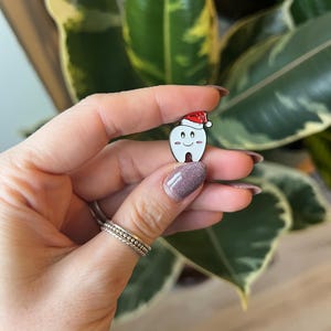Pu&ograve; includere: Una spilla di smalto bianco a forma di dente con un cappello da Babbo Natale rosso. Il dente ha una faccia sorridente con occhi neri e bocca rosa. La spilla &egrave; tenuta davanti a uno sfondo di foglie verdi.