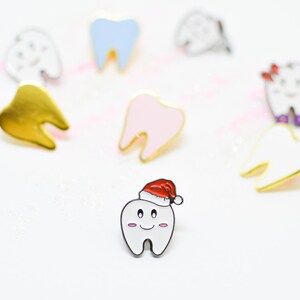 Pu&ograve; includere: Una collezione di spille smaltate a forma di denti dei cartoni animati. Le spille sono in vari colori, tra cui oro, bianco, rosa e blu. Una spilla a forma di dente indossa un cappello di Babbo Natale. Le spille hanno contorni neri su sfondo bianco.