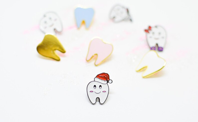 Pu&ograve; includere: Una collezione di spille smaltate a forma di denti dei cartoni animati. Le spille sono in vari colori, tra cui oro, bianco, rosa e blu. Una spilla a forma di dente indossa un cappello di Babbo Natale. Le spille hanno contorni neri su sfondo bianco.
