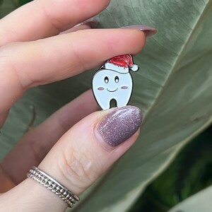 Pu&ograve; includere: Una spilla di smalto bianco a forma di dente, con un cappello da Babbo Natale rosso. Il dente ha una faccia sorridente con guance rosee. La spilla &egrave; tenuta su uno sfondo di foglie verdi.