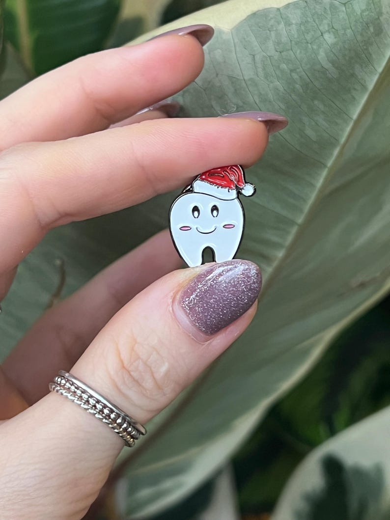 Pu&ograve; includere: Una spilla di smalto bianco a forma di dente, con un cappello da Babbo Natale rosso. Il dente ha una faccia sorridente con guance rosee. La spilla &egrave; tenuta su uno sfondo di foglie verdi.