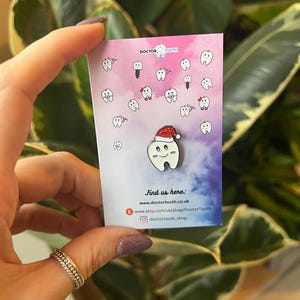 Pu&ograve; includere: Spilla in smalto bianco raffigurante un dente sorridente con un cappello da Babbo Natale, su una carta con illustrazioni di denti e la scritta "Doctor Tooth". Lo sfondo &egrave; sfumato rosa e viola.