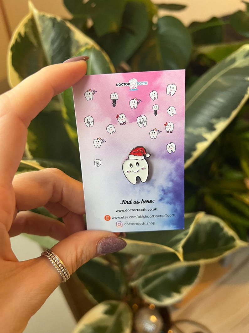 Pu&ograve; includere: Spilla in smalto bianco raffigurante un dente sorridente con un cappello da Babbo Natale, su una carta con illustrazioni di denti e la scritta "Doctor Tooth". Lo sfondo &egrave; sfumato rosa e viola.