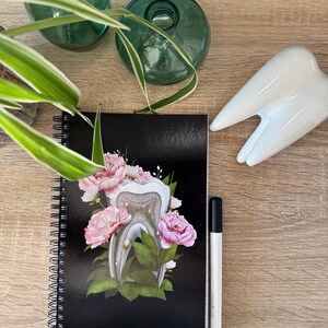 K&ouml;nnte beinhalten: Ein schwarzes Spiralnotizbuch mit einem wei&szlig;en Zahn, umgeben von rosa Blumen und gr&uuml;nen Bl&auml;ttern. Das Notizbuch liegt auf einer Holzoberfl&auml;che mit einem wei&szlig;en Zahn und einem schwarzen Stift.