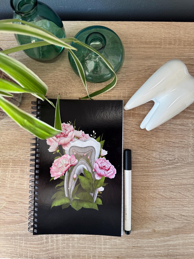 K&ouml;nnte beinhalten: Ein schwarzes Spiralnotizbuch mit einem wei&szlig;en Zahn, umgeben von rosa Blumen und gr&uuml;nen Bl&auml;ttern. Das Notizbuch liegt auf einer Holzoberfl&auml;che mit einem wei&szlig;en Zahn und einem schwarzen Stift.