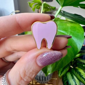 May include: A lilac enamel tooth-shaped pin with a gold-coloured outline. The pin is held in front of a green leafy plant. The pin is a cute accessory for dental professionals or anyone who loves teeth.