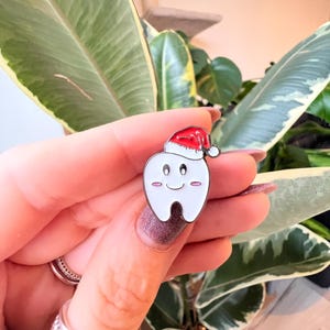 Pu&ograve; includere: Una spilla di smalto bianco a forma di dente, con un cappello da Babbo Natale rosso. Il dente ha una faccia sorridente con guance rosa. La spilla &egrave; tenuta davanti a uno sfondo di foglie verdi e bianche.