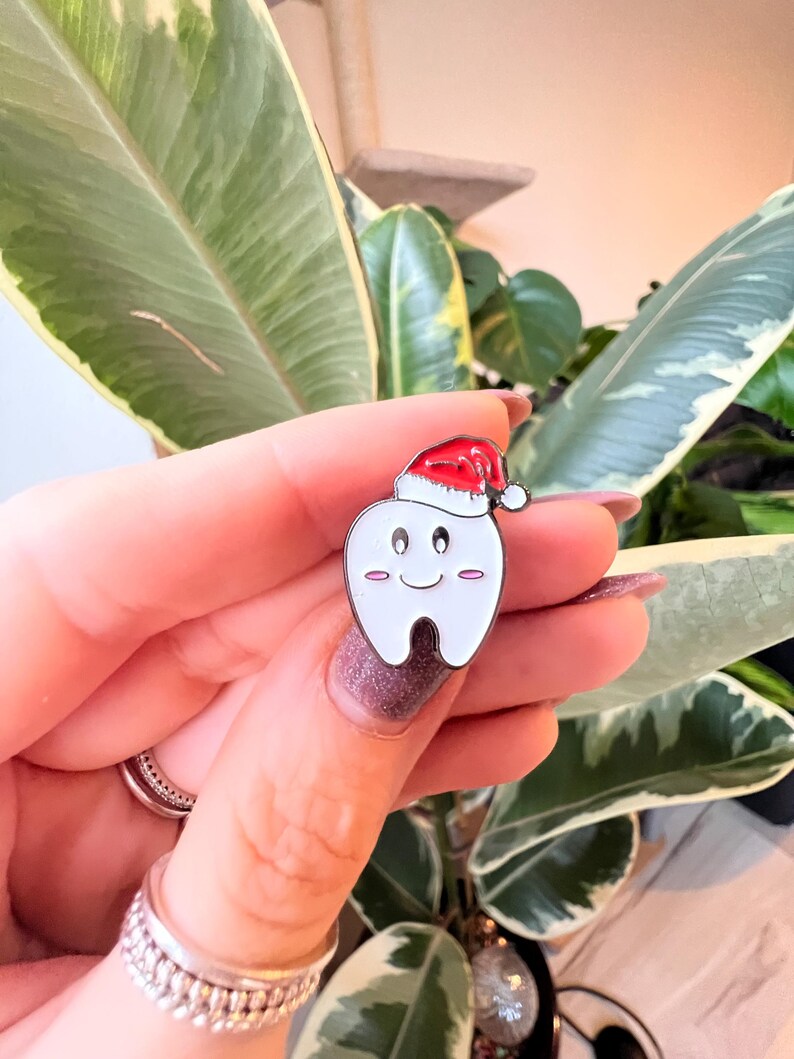 Pu&ograve; includere: Una spilla di smalto bianco a forma di dente, con un cappello da Babbo Natale rosso. Il dente ha una faccia sorridente con guance rosa. La spilla &egrave; tenuta davanti a uno sfondo di foglie verdi e bianche.