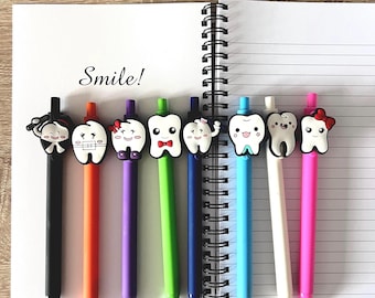 May include: A collection of eight colorful pens with tooth-shaped toppers, each with a unique design. The pens are arranged on a spiral-bound notebook, with the word "Smile!" written above them. The pens have black, orange, purple, green, blue, white, and pink barrels.