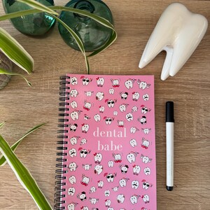 K&ouml;nnte beinhalten: Rosa gebundenes Notizbuch mit einem wei&szlig;en Zahnmuster und dem Text "dental babe" auf dem Cover. Das Notizbuch liegt auf einer Holzoberfl&auml;che mit einem wei&szlig;en Keramikzahn.