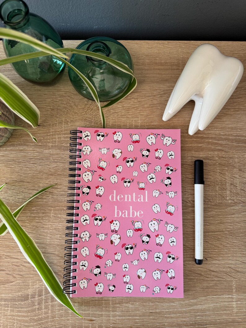 K&ouml;nnte beinhalten: Rosa gebundenes Notizbuch mit einem wei&szlig;en Zahnmuster und dem Text "dental babe" auf dem Cover. Das Notizbuch liegt auf einer Holzoberfl&auml;che mit einem wei&szlig;en Keramikzahn.
