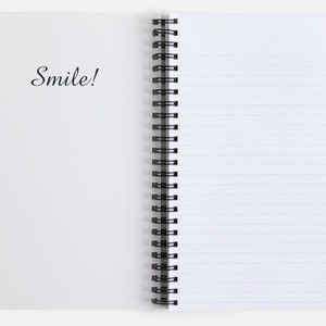 K&ouml;nnte beinhalten: Ein wei&szlig;es Spiralblock mit linierten Seiten. Der Einband hat den Schriftzug "Smile!" in schwarzer Kursivschrift.