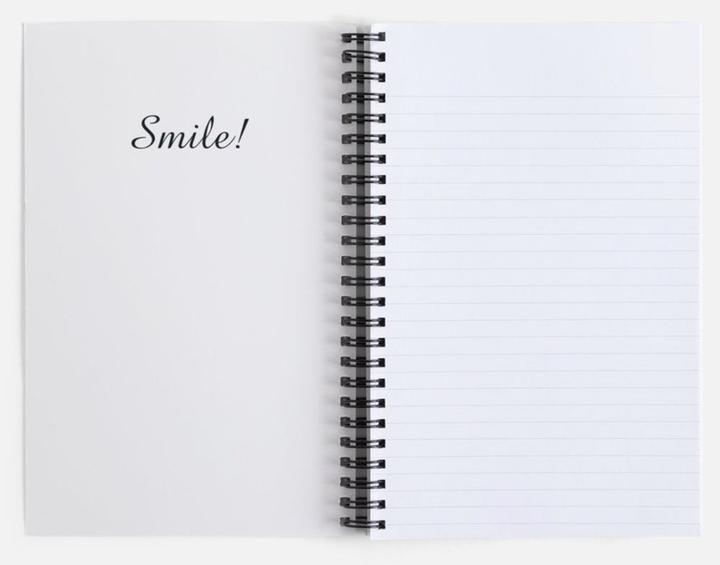 K&ouml;nnte beinhalten: Ein wei&szlig;es Spiralblock mit linierten Seiten. Der Einband hat den Schriftzug "Smile!" in schwarzer Kursivschrift.