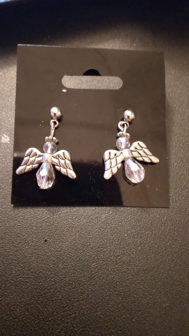 May include: A pair of silver angel earrings with clear crystal accents. The earrings are on a black card.