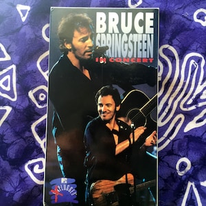 Puede incluir: Una cinta VHS con Bruce Springsteen en concierto. La portada muestra a Springsteen tocando la guitarra, con el texto "BRUCE SPRINGSTEEN IN CONCERT" en blanco y rojo. El logotipo de MTV "Plugged" es visible.