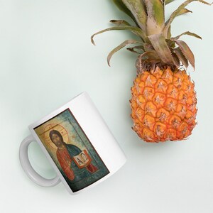 May include: White ceramic mug featuring a religious icon of Jesus Christ holding a book. The mug has a curved handle and a rectangular image. A ripe pineapple with green leaves sits nearby on a light blue background.