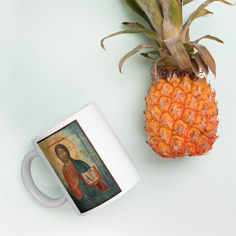May include: White ceramic mug featuring a religious icon of Jesus Christ holding a book. The mug has a curved handle and a rectangular image. A ripe pineapple with green leaves sits nearby on a light blue background.