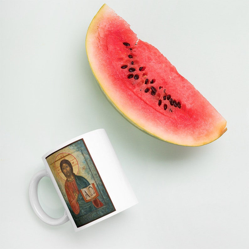 May include: A white ceramic mug featuring a religious icon with a halo, red robe, and open book. A large slice of watermelon with black seeds is next to the mug. The watermelon has a green rind and bright red flesh.