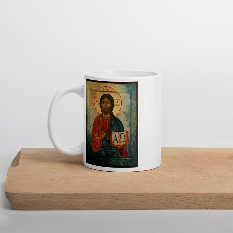 May include: White ceramic mug featuring a religious icon. The icon depicts a figure in red and blue robes, holding a book with Greek letters. The background is a gold halo. The mug is on a wooden surface.