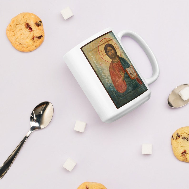 May include: White ceramic mug featuring a religious icon illustration. The artwork depicts a figure with a halo, wearing red and blue robes, holding a book. The mug has a curved handle and is surrounded by cookies, sugar cubes, and a spoon.