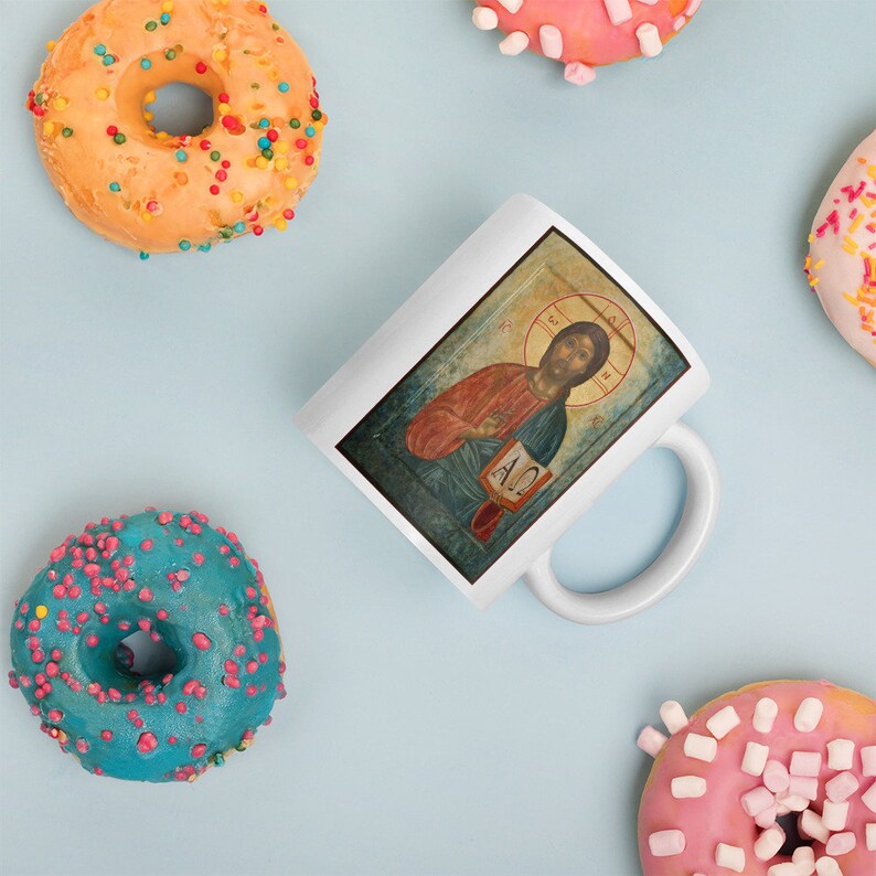 May include: White ceramic mug featuring a religious icon of Jesus Christ, with a red and gold color scheme. The mug is surrounded by colorful donuts with various toppings, including sprinkles and marshmallows. The background is a light blue.